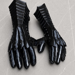 K&ouml;nnte beinhalten: Ein Paar schwarze, bewegliche Metallhandschuhe mit &uuml;berlappenden Platten, die Finger, H&auml;nde und Handgelenke bedecken. Die R&uuml;stungsteile sind durch kleine Nieten verbunden und haben eine gl&auml;nzende Oberfl&auml;che f&uuml;r einen vollst&auml;ndigen Handschutz.