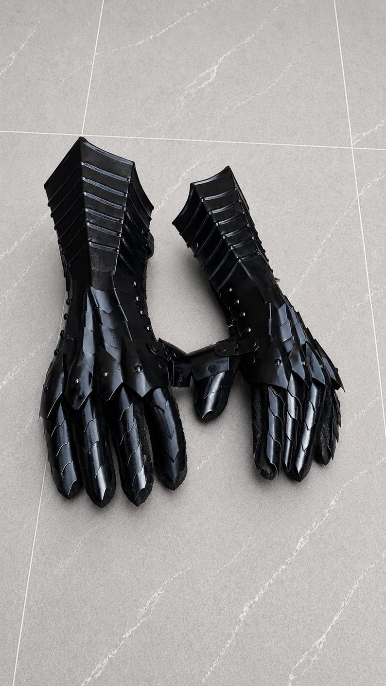 K&ouml;nnte beinhalten: Ein Paar schwarze, bewegliche Metallhandschuhe mit &uuml;berlappenden Platten, die Finger, H&auml;nde und Handgelenke bedecken. Die R&uuml;stungsteile sind durch kleine Nieten verbunden und haben eine gl&auml;nzende Oberfl&auml;che f&uuml;r einen vollst&auml;ndigen Handschutz.