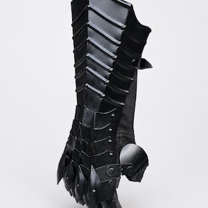 K&ouml;nnte beinhalten: Ein schwarzer, beweglicher Panzerhandschuh mit geschichteten Metallplatten, die Unterarm und Hand bedecken. Das Design umfasst eine Reihe &uuml;berlappender Segmente, die mit Nieten befestigt sind und vom Handgelenk bis zu den Kn&ouml;cheln reichen.