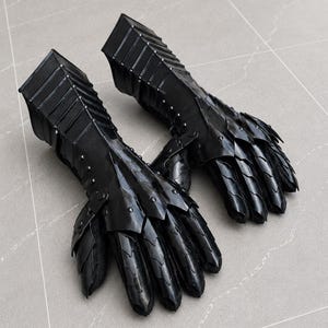 K&ouml;nnte beinhalten: Ein Paar schwarze, bewegliche Panzerhandschuhe mit geschichteten Metallplatten, die die Finger und den Handr&uuml;cken bedecken und mit einer starren Unterarmmanschette mit sichtbaren Nieten verbunden sind. Das Design umfasst &uuml;berlappende Segmente.