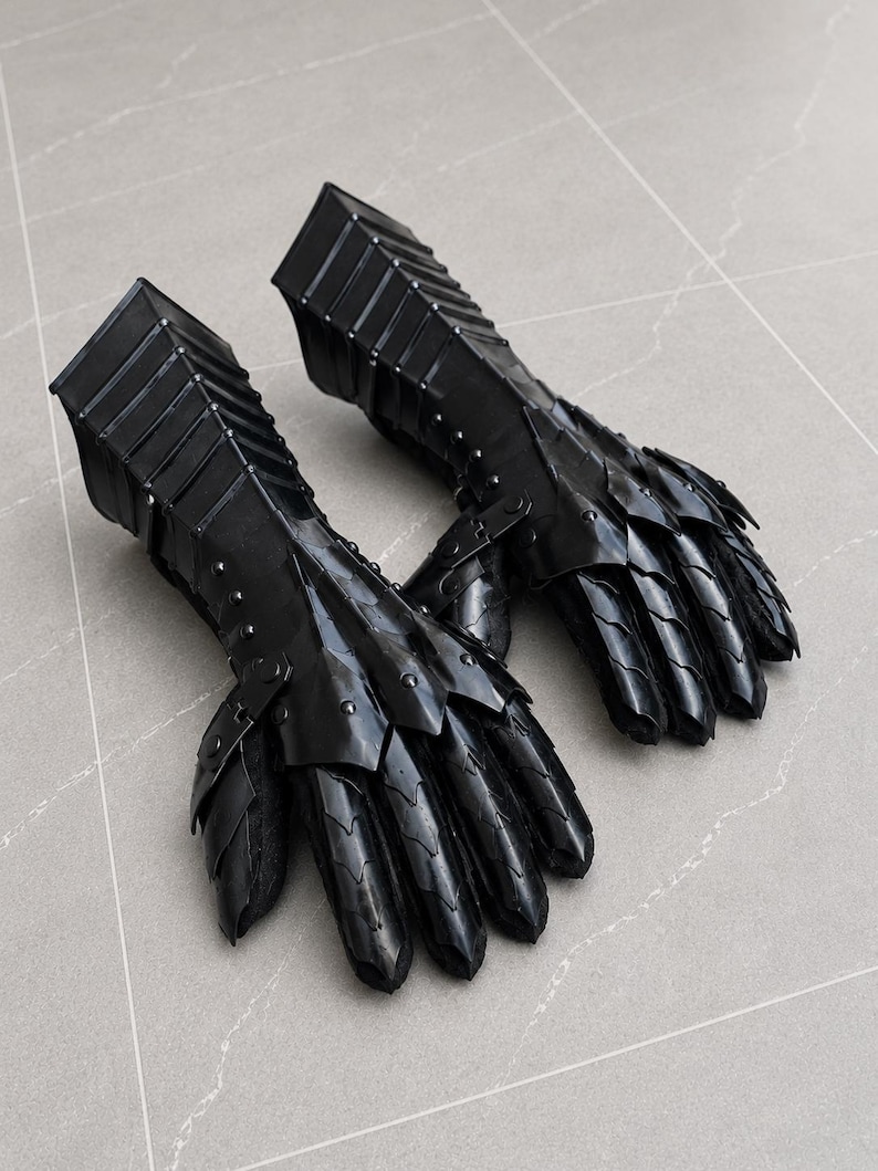 K&ouml;nnte beinhalten: Ein Paar schwarze, bewegliche Panzerhandschuhe mit geschichteten Metallplatten, die die Finger und den Handr&uuml;cken bedecken und mit einer starren Unterarmmanschette mit sichtbaren Nieten verbunden sind. Das Design umfasst &uuml;berlappende Segmente.