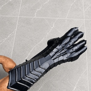 K&ouml;nnte beinhalten: Ein schwarzer, beweglicher Panzerhandschuh mit segmentiertem Design, der den Unterarm bedeckt und &uuml;ber die Finger reicht. Das R&uuml;stungsteil besteht aus mehreren &uuml;berlappenden Platten, die durch Nieten verbunden sind und eine flexible Schutzstruktur bilden.