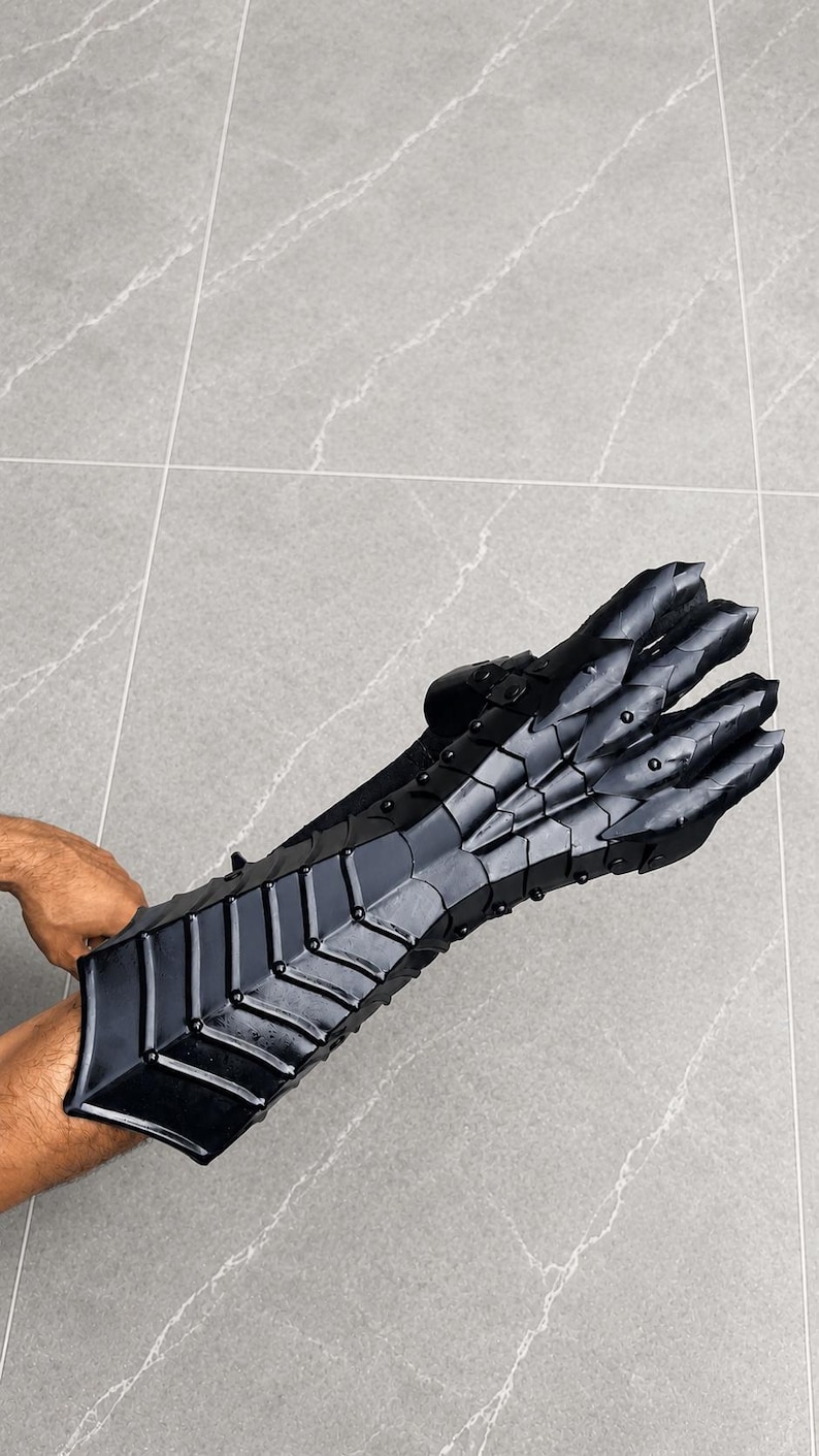 K&ouml;nnte beinhalten: Ein schwarzer, beweglicher Panzerhandschuh mit segmentiertem Design, der den Unterarm bedeckt und &uuml;ber die Finger reicht. Das R&uuml;stungsteil besteht aus mehreren &uuml;berlappenden Platten, die durch Nieten verbunden sind und eine flexible Schutzstruktur bilden.