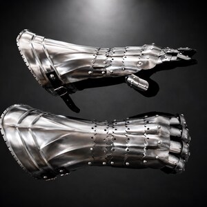 Könnte beinhalten: Ein Paar bewegliche Metallhandschuhe mit polierter silberner Oberfläche. Jeder Handschuh besteht aus überlappenden, mit Nieten befestigten Stahlplatten, die vom Handgelenk bis zu den Fingern reichen, mit einem schwarzen Lederriemen und Schnalle.