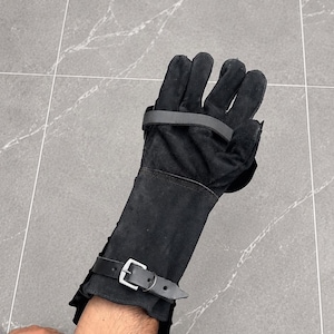 K&ouml;nnte beinhalten: Ein schwarzer Wildleder-Handschuh mit langem Schaft, der den Unterarm bedeckt. Der Handschuh verf&uuml;gt &uuml;ber einen horizontalen Riemen &uuml;ber dem Handr&uuml;cken und einen verstellbaren Schnallenriemen am Handgelenk, beide aus passendem schwarzem Material.
