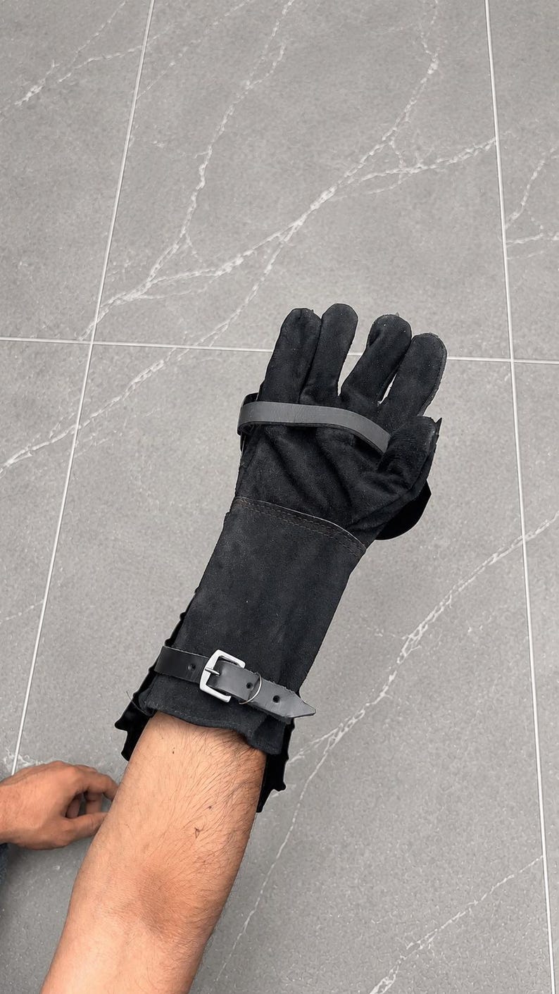 K&ouml;nnte beinhalten: Ein schwarzer Wildleder-Handschuh mit langem Schaft, der den Unterarm bedeckt. Der Handschuh verf&uuml;gt &uuml;ber einen horizontalen Riemen &uuml;ber dem Handr&uuml;cken und einen verstellbaren Schnallenriemen am Handgelenk, beide aus passendem schwarzem Material.