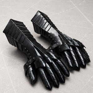 K&ouml;nnte beinhalten: Ein Paar schwarze Metallhandschuhe mit beweglichen Fingerplatten und einer starren Unterarmmanschette. Die R&uuml;stungsteile sind mit kleinen Nieten und Scharnieren verbunden, wodurch ein geschichtetes, segmentiertes Design entsteht, das Hand und Handgelenk bedeckt.