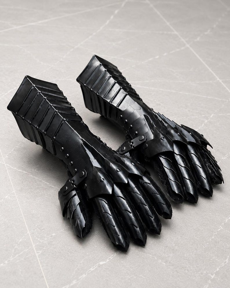 K&ouml;nnte beinhalten: Ein Paar schwarze Metallhandschuhe mit beweglichen Fingerplatten und einer starren Unterarmmanschette. Die R&uuml;stungsteile sind mit kleinen Nieten und Scharnieren verbunden, wodurch ein geschichtetes, segmentiertes Design entsteht, das Hand und Handgelenk bedeckt.