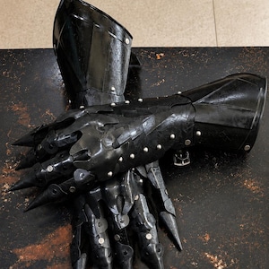 Könnte beinhalten: Ein Paar schwarze Panzerhandschuhe mit beweglichen Fingerplatten mit spitzen Enden, geschichteten Metallplatten an Hand- und Handgelenksbereichen sowie kleinen silberfarbenen Nieten, die die einzelnen Komponenten zusammenhalten.