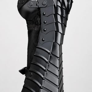 K&ouml;nnte beinhalten: Ein schwarzer, beweglicher Panzerhandschuh mit &uuml;berlappenden Metallplatten, die mit Nieten befestigt sind. Das R&uuml;stungsteil bedeckt Unterarm und Hand, mit einem separaten Daumenschutz und einem dunklen Stofffutter unter der Metallstruktur.