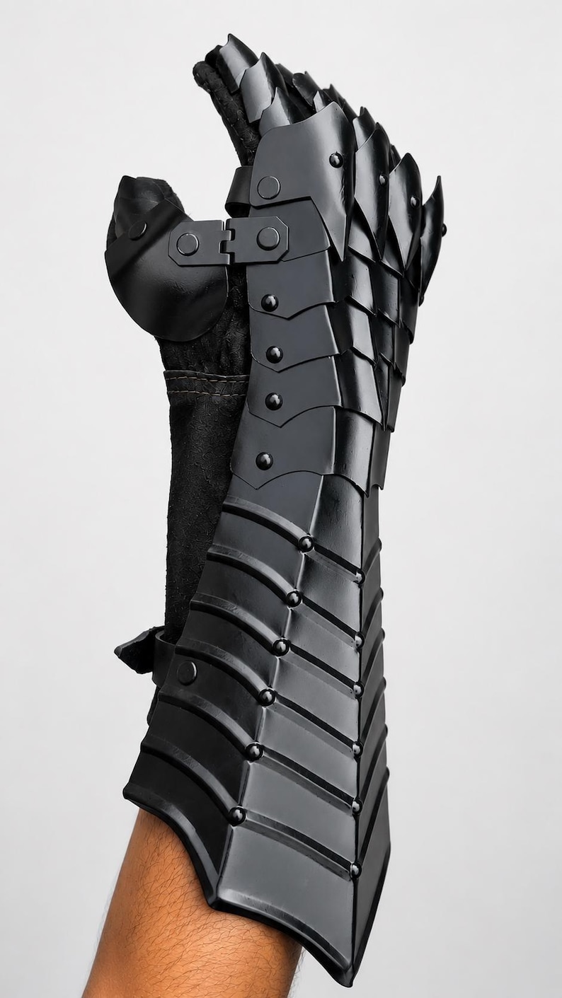 K&ouml;nnte beinhalten: Ein schwarzer, beweglicher Panzerhandschuh mit &uuml;berlappenden Metallplatten, die mit Nieten befestigt sind. Das R&uuml;stungsteil bedeckt Unterarm und Hand, mit einem separaten Daumenschutz und einem dunklen Stofffutter unter der Metallstruktur.
