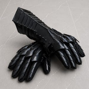 K&ouml;nnte beinhalten: Ein Paar schwarze, bewegliche Panzerhandschuhe mit &uuml;berlappenden Metallplatten, die die Finger und den Handr&uuml;cken bedecken, verbunden mit einer starren, segmentierten Manschette, die den Unterarm bedeckt und mit Nieten befestigt ist.