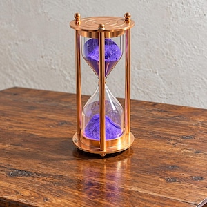 May include: An hourglass featuring a metallic copper frame with circular top and bottom plates supported by four vertical rods. The clear glass bulb contains vibrant purple sand, with a portion currently resting in the bottom chamber.