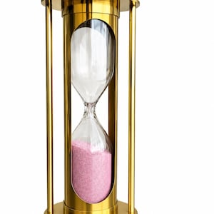 Op de afbeelding: Een goudkleurige metalen zandloper met een cilindrisch frame en verticale steunpilaren. De centrale glazen houder bevat roze zand, waarbij het grootste deel van het zand zich in de onderste bol bevindt en een kleine hoeveelheid in de bovenste bol.