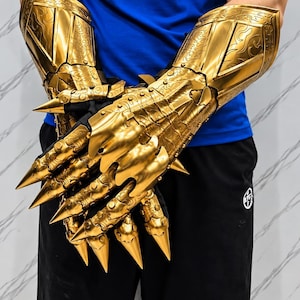 Könnte beinhalten: Ein Paar goldfarbene Panzerhandschuhe mit detaillierten, geprägten Schnörkelmustern und langen, spitzen, krallenartigen Fingerverlängerungen. Die metallischen Rüstungsteile bedecken Hände und Unterarme und weisen eine polierte Oberfläche mit scharfen, konischen Spitzen auf.
