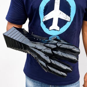 K&ouml;nnte beinhalten: Ein schwarzer, beweglicher Metallhandschuh mit &uuml;berlappenden Platten, die Hand und Unterarm bedecken. Der Handschuh hat eine gl&auml;nzende Oberfl&auml;che und ist aus mehreren Segmenten gefertigt, die Beweglichkeit erm&ouml;glichen, mit einem detaillierten Design.