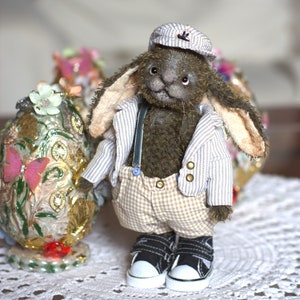 May include: A plush bunny wearing a striped shirt, plaid pants, a cap, and sneakers. The bunny is standing on a white lace tablecloth next to a decorative egg.