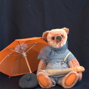 May include: A brown teddy bear wearing a blue and white striped shirt, a blue scarf, and overalls. The bear is holding a brown paper bag and sitting under an orange umbrella. The bear is wearing a gray beret.