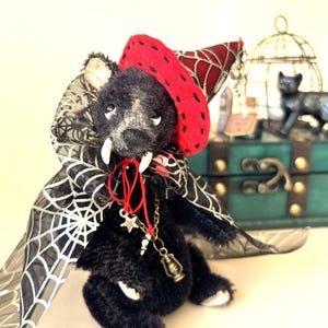 Peut inclure: Un ours en peluche noir portant un chapeau pointu rouge à motifs sombres, une cape noire transparente avec un imprimé toile d'araignée blanche et deux crocs blancs. L'ours porte un collier doré avec une étoile et une lanterne miniature.