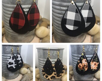 May include: Four pairs of dangle earrings with a teardrop shape. Each pair features a different pattern: red and black buffalo plaid, black and white buffalo plaid, leopard print, and floral print. The earrings are made of faux leather and have gold-toned hooks.