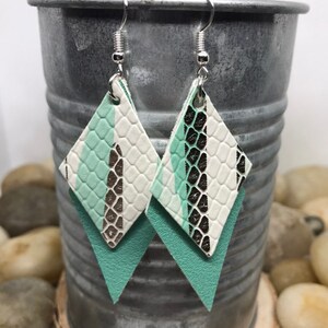 May include: A pair of dangle earrings with a layered design. The top layer is white with a snakeskin pattern and a silver stripe. The bottom layer is a solid mint green. The earrings are attached to silver hooks.