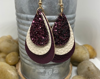 May include: A pair of dangle earrings with three layers of leather. The top layer is burgundy glitter, the middle layer is cream, and the bottom layer is burgundy. The earrings have gold hooks.