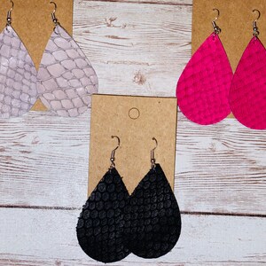 May include: Three pairs of teardrop-shaped leather earrings. The earrings are in white, pink, and black. Each pair has a different pattern.