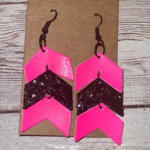 May include: A pair of pink and black chevron earrings with glitter accents. The earrings are made of leather and have a simple hook closure.