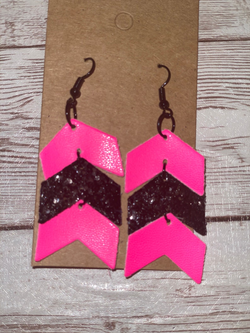 May include: A pair of pink and black chevron earrings with glitter accents. The earrings are made of leather and have a simple hook closure.