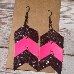 May include: A pair of black and pink chevron glitter earrings. The earrings are made of leather and have a black glitter finish with a pink chevron pattern.