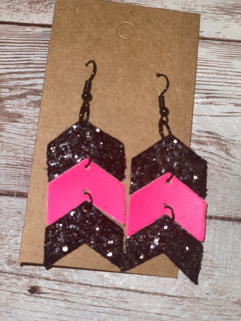 May include: A pair of black and pink chevron glitter earrings. The earrings are made of leather and have a black glitter finish with a pink chevron pattern.