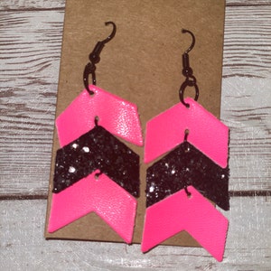 May include: A pair of pink and black chevron earrings with glitter accents. The earrings are made of leather and have a simple hook closure.