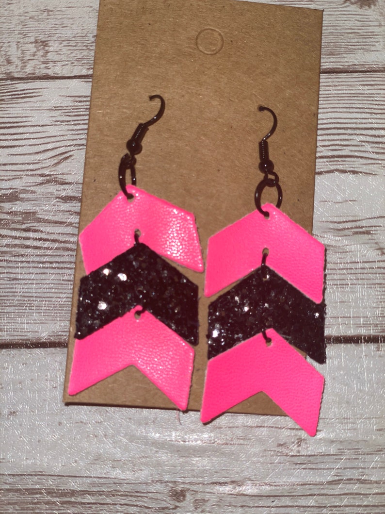 May include: A pair of pink and black chevron earrings with glitter accents. The earrings are made of leather and have a simple hook closure.