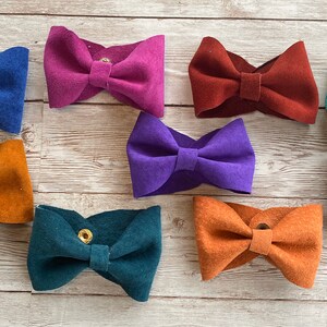 May include: A collection of suede bow ties in various colors, including blue, purple, orange, and teal. Each bow tie is secured with a snap closure. The bow ties are arranged on a light wood-grain surface.