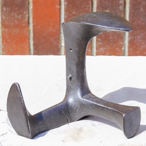 May include: A vintage, dark grey cast iron cobbler's last for shaping shoes. The tool features a curved top, a central support, and a three-pronged base. The surface shows signs of wear, suggesting its age and use in shoemaking.