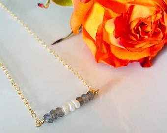May include: A delicate gold chain necklace featuring a bar pendant with a row of white pearls and gray-blue labradorite beads. An orange rose is in the background, adding a touch of color to the jewelry.