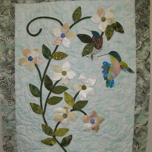 May include: A blue and green hummingbird with a long beak perched on a branch of white flowers with green leaves against a light blue background.