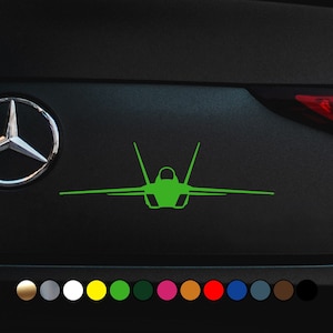 May include: Green silhouette of a fighter jet on a black car. The silhouette is facing forward and has a sharp nose and wings. The car has a silver front bumper and a black grille.