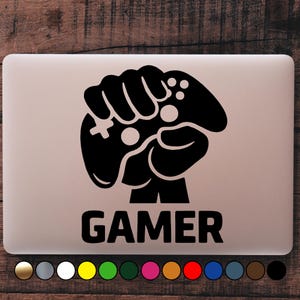 Può includere: Grafica nera di una mano che tiene un controller di videogiochi, con la parola "GAMER" sotto. Il design è su un laptop, con una selezione di opzioni di colore in basso.