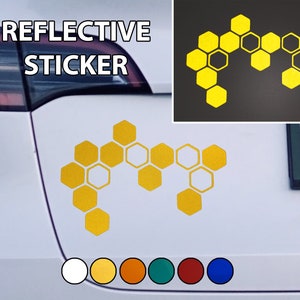 Op de afbeelding: Een reflecterende sticker met een honingraatontwerp in geel. De sticker zit op een witte auto. De tekst "REFLECTIVE STICKER" staat boven de sticker.