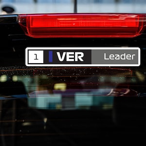 以下が含まれることがあります： 車の窓に貼られた黒と白のステッカーに「1 VER Leader」と書かれている。
