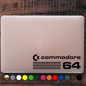 Può includere: Un laptop con un adesivo nero Commodore 64. L'adesivo presenta il logo Commodore, la parola "commodore", linee orizzontali e il numero "64". Il laptop è di colore beige chiaro.