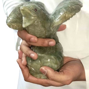 Könnte beinhalten: Eine grüne Jade-Schnitzerei eines Vogels mit ausgebreiteten Flügeln. Der Vogel hat ein rotes Auge und wird in den Händen einer Person gehalten.