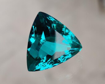 May include: A triangular, faceted gemstone with a vibrant teal color. The gem's multiple facets reflect light, creating a sparkling effect. The gemstone is set against a neutral background, highlighting its color and shape.