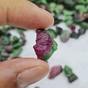 May include: A rough ruby gemstone with a deep red hue, held between fingers. The ruby has a textured surface with green edges. Other raw gemstones are scattered in the background.