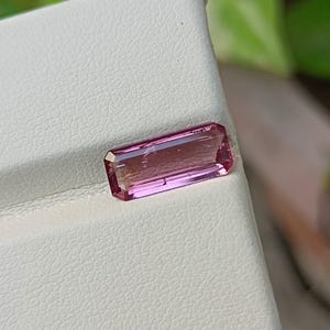 May include: A rectangular, faceted pink gemstone with a step-cut design. The stone displays a gradient of color, transitioning from a deeper magenta at the edges to a lighter, translucent pink in the center, reflecting light across its surface.