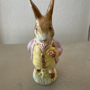 May include: A brown and white ceramic figurine of a rabbit wearing a pink jacket, yellow vest, and a red and white striped tie. The rabbit is holding a small brown object in its right paw.