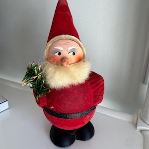 May include: A vintage Christmas gnome figurine. The gnome wears a red velvet hat and suit with a white fur beard and collar. The gnome holds a small green sprig and has black shoes.