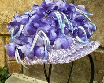 May include: A vintage hat with a lavender brim and a crown of layered purple fabric flowers. White and light blue ribbons cascade from the floral arrangement. The hat is displayed on a black metal stand, showcasing its intricate design.