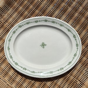 May include: White oval serving platter with a green floral border and a small floral design in the center. The platter is made of ceramic and is set on a woven wicker surface.